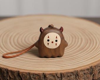 Op de afbeelding: Een kleine, uit hout gesneden figuurhanger met een glad, donkerbruin lichaam en twee kleine hoorntjes. Het heeft een lichtgekleurd, rond gezichtsinleg met twee zwarte ogen, een kleine rode stip en vier rode driehoekige markeringen aan de zijkanten.