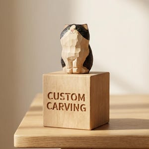 May include: A wooden cat carving with black, white, and tan markings, sitting on a wooden cube. The cube is engraved with the words "CUSTOM CARVING". The cat sculpture is detailed and has a whimsical appearance.