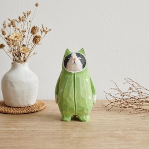 May include: A hand-carved wooden cat figurine wearing a green jumpsuit with white star accents. The cat has a black and white face and is standing upright. The figurine is on a wooden surface.