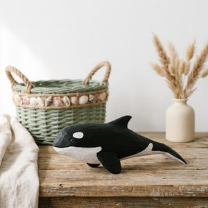 Puede incluir: Una figura de orca tallada a mano en madera con un acabado liso en blanco y negro y marcas de cincel visibles. La ballena se muestra en posición de nado horizontal con una aleta dorsal prominente, aletas laterales y una mancha blanca cerca del ojo.