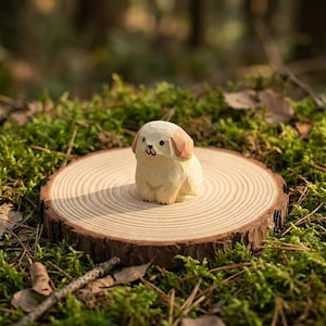 May include: A hand-carved wooden dog figurine with a light beige body and brown ears, placed on a small, round wooden slice. The dog features a simple, stylized design with black eyes and nose. It is set against a backdrop of green moss.