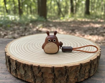 Op de afbeelding: Een bruin houten Totoro-beeldje met een lichtbruine buik en grote, ronde ogen. Het beeldje heeft een bruin leren bandje. Het beeldje staat op een houten schijf met een natuurlijke schorsrand.