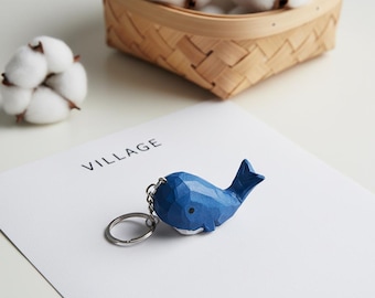 Op de afbeelding: Een blauwe walvis sleutelhanger met een zilveren sleutelring. De walvis is gesneden met geometrische facetten en heeft een witte buik. Het woord "VILLAGE" staat op het witte papier onder de sleutelhanger. Een mand met wattenbollen staat op de achtergrond.