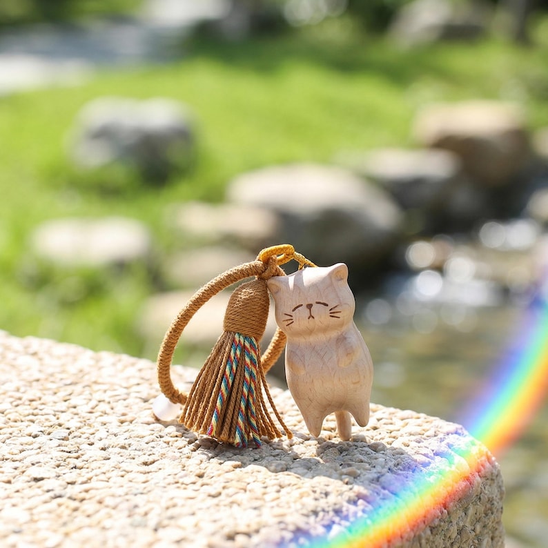 Op de afbeelding: Een houten kattenbeeldje met een kwastjes sleutelhanger. De kat is gesneden uit lichtgekleurd hout en heeft eenvoudige gelaatstrekken. De kwast is bruin met kleurrijke draden. De achtergrond is wazig en toont groen en een regenboog.