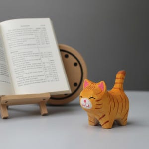 May include: A wooden cat figurine, hand-carved with orange and brown stripes, pink ears, and a smiling face. The cat is positioned near an open book on a wooden easel and a circular decorative object.
