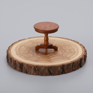 Puede incluir: Una mesa de madera en miniatura con una tapa redonda, de pie sobre una rodaja de madera natural. La mesa es de color marrón oscuro, y la rodaja de madera tiene un interior marrón claro con un borde de corteza marrón oscuro. La mesa mide unos 5 cm de alto.