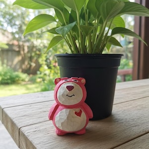 Könnte beinhalten: Handgeschnitzte Holzfigur eines rosa-weißen Bären mit Herz. Der Bär steht neben einer Topfpflanze in einem schwarzen Topf auf einer Holzoberfläche.