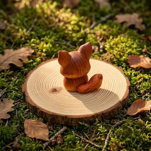 K&ouml;nnte beinhalten: Eine kleine, glatte, rotbraune Holzfigur eines Fuchses mit spitzen Ohren und einem geschwungenen Schwanz, platziert auf einer runden, hellen Holzscheibe mit sichtbaren Jahresringen und einer nat&uuml;rlichen Rindenkante.