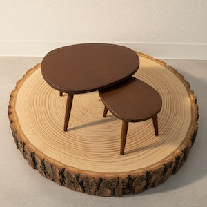 May include: Two brown, organic-shaped side tables with three wooden legs each. The tables are nested together, with the smaller table underneath the larger one. The tables are set on a large, round, light brown wood slice.