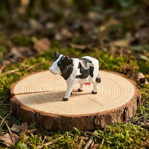 Puede incluir: Figura de vaca de madera tallada a mano, con marcas en blanco y negro, sobre una rodaja de madera natural. La vaca tiene una ubre roja y patas marrones. La rodaja de madera está rodeada de musgo y hojas verdes.