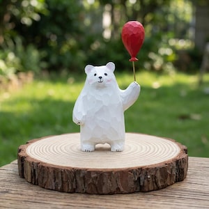 Pode incluir: Uma figura de urso branco de madeira segurando um balão vermelho. O urso tem uma textura esculpida e bochechas rosadas. Ele está em uma fatia de madeira redonda com borda de casca natural. O fundo é verde desfocado.