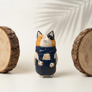 Puede incluir: Figura de gato de madera tallada a mano con cabeza tricolor naranja, negra y blanca. El gato lleva una bufanda azul con lunares blancos y un peto azul con lunares marrones. Dos rodajas de madera flanquean al gato sobre un fondo blanco.
