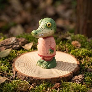 Könnte beinhalten: Eine skurrile grüne Alligatorfigur mit großen gelben Augen, die einen rosa Pullover und grüne Hosen trägt. Die Figur steht auf einer Holzscheibe, umgeben von Moos und Blättern, wodurch eine natürliche, naturverbundene Szene entsteht.