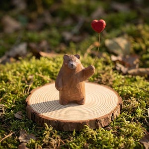 May include: A carved wooden bear figurine holding a red heart-shaped balloon. The bear stands on a small, round wooden slice, set against green moss. The figurine has a rustic, hand-crafted appearance.