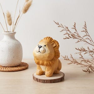 May include: A small, hand-carved wooden lion figurine with a light brown finish and a textured mane. The lion is seated on a circular, natural wood slice base with visible bark edges, showcasing detailed wood grain and carved facial features.