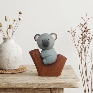 Könnte beinhalten: Eine handgeschnitzte Koala-Figur aus Holz mit glatter grauer Oberfläche, abgerundeten Ohren und einer dunklen Nase, die auf einem Sockel aus einem natürlichen braunen Holzast sitzt. Der Koala hat eine facettierte, skulpturale Optik.