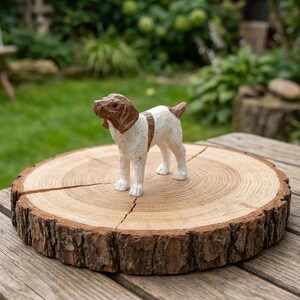 May include: A hand-carved, white and brown dog figurine stands on a rustic wooden slice. The dog has brown ears and a brown spot over its eyes. The wooden slice has a natural bark edge and is placed on a wooden surface. The background is a blurred green garden.