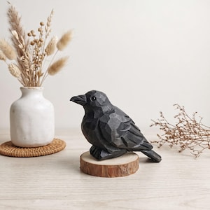 Puede incluir: Una figura de cuervo de madera tallada en color negro con una textura de superficie facetada, posada sobre una pequeña base circular de rodaja de madera clara. El ave tiene un pico oscuro y un ojo liso y redondeado, con un acabado detallado.