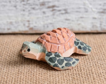 May include: A small hand-carved wooden sea turtle figurine featuring a light brown shell with visible carved facets, a light tan head, and light green flippers decorated with dark green spots. The turtle is shown in a side profile view.