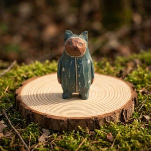 Op de afbeelding: Handgesneden houten kattenfiguur in een blauwe onesie met witte sterren. De kat staat op een ronde houten schijf, tegen een achtergrond van groen mos en bosbladeren. Het figuur heeft een grillige, volkse stijl.
