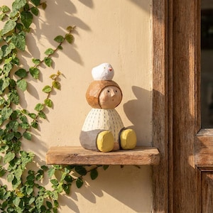 May include: A wooden figurine of a person with a bird on its head. The figurine has a brown head, a cream body with vertical lines, grey trousers, and yellow feet. It sits on a wooden shelf against a beige wall. The shelf is made of wood.