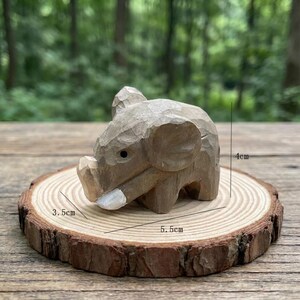 K&ouml;nnte beinhalten: Eine kleine, handgeschnitzte Holz-Elefantenfigur, ca. 4 cm hoch, 3,5 cm breit und 5,5 cm lang. Der Elefant ist hellbraun und sitzt auf einer Holzscheibe mit nat&uuml;rlichem Rand. Der Hintergrund ist unscharf.
