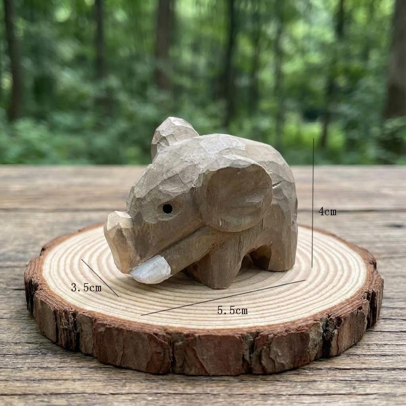 K&ouml;nnte beinhalten: Eine kleine, handgeschnitzte Holz-Elefantenfigur, ca. 4 cm hoch, 3,5 cm breit und 5,5 cm lang. Der Elefant ist hellbraun und sitzt auf einer Holzscheibe mit nat&uuml;rlichem Rand. Der Hintergrund ist unscharf.