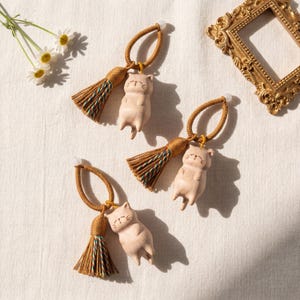 Op de afbeelding: Drie beige katvormige ornamenten met bruine kwastjes en lussen. De katfiguren liggen met gesloten ogen. De kwastjes hebben groene en blauwe accenten. De ornamenten liggen op een wit oppervlak met een gouden lijst en bloemen.