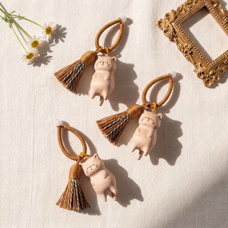 Op de afbeelding: Drie beige katvormige ornamenten met bruine kwastjes en lussen. De katfiguren liggen met gesloten ogen. De kwastjes hebben groene en blauwe accenten. De ornamenten liggen op een wit oppervlak met een gouden lijst en bloemen.