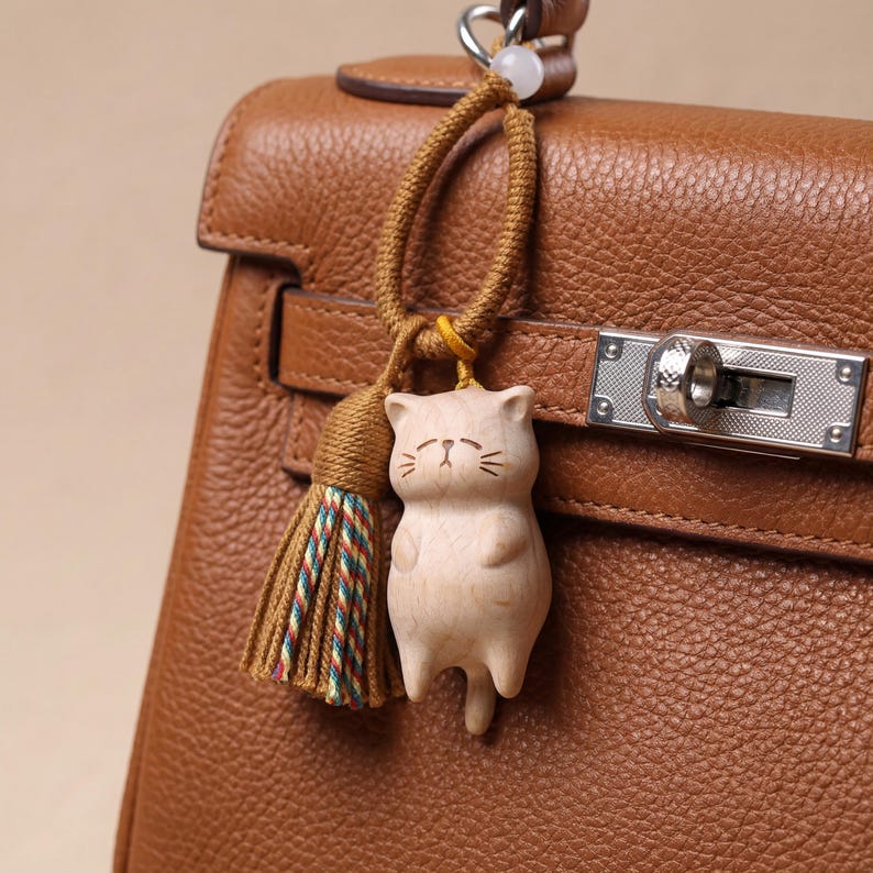 Op de afbeelding: Een houten kattenbedel met een kwast, hangend aan een bruine leren handtas. De kattenbedel is licht van kleur met ge&euml;tste gezichtskenmerken. De kwast heeft bruine, groene en rode draden. De handtas is een middellange bruine kleur.