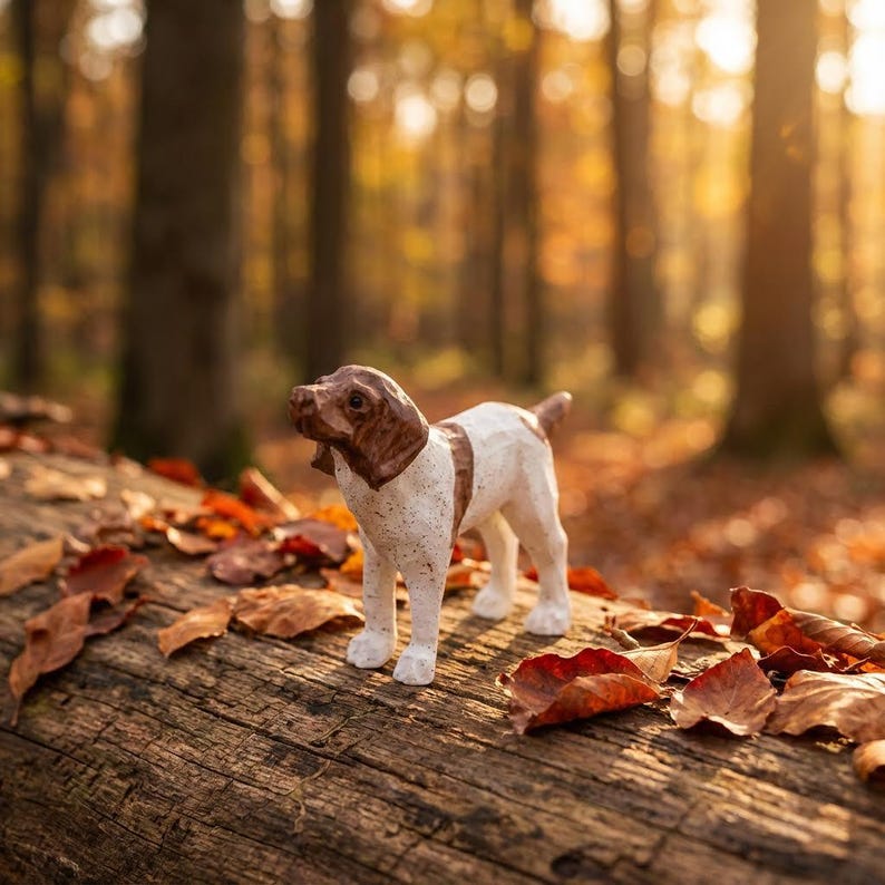 May include: A small, handcrafted dog figurine with a brown and white speckled coat stands on a weathered log. The dog's head and back are brown, while the rest of its body is white. The background features a blurred forest with autumn leaves.