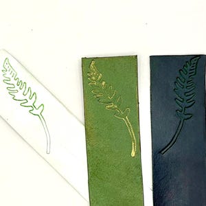 May include: Three rectangular bookmarks with a fern design. One is white with a green outline, one is green with a gold design, and the last is dark blue with a debossed design. The bookmarks are arranged on a white background.