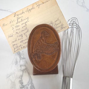 May include: A wooden duck-shaped butter mold with a recipe card for "French Apple Squares" and a metal whisk. The recipe card is handwritten. The butter mold is oval and stands upright. The whisk has a silver handle.