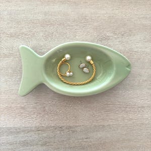 May include: A light green ceramic fish-shaped trinket dish holding gold jewelry with pearls. The dish has a tail and a head with a small opening. The jewelry includes a gold bracelet, a ring, and a pair of earrings.