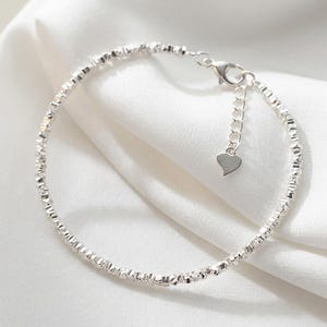 May include: A delicate silver-toned bracelet featuring a textured, faceted chain design. The bracelet includes a thin extender chain finished with a small, flat heart-shaped charm and a secure lobster claw clasp closure mechanism.