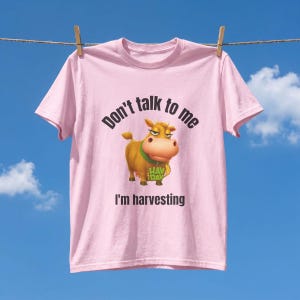 May include: A light pink short-sleeved t-shirt featuring a graphic of a brown cow with a green collar and the Hay Day logo. The text above the cow reads Don't talk to me and the text below the cow reads I'm harvesting in black lettering.