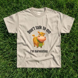 May include: A beige short-sleeved t-shirt featuring a graphic of a brown cow with a green collar. The text above the cow reads "Don't talk to me" and the text below reads "I'm harvesting". The cow also has the word "HAY DAY" printed on its chest.