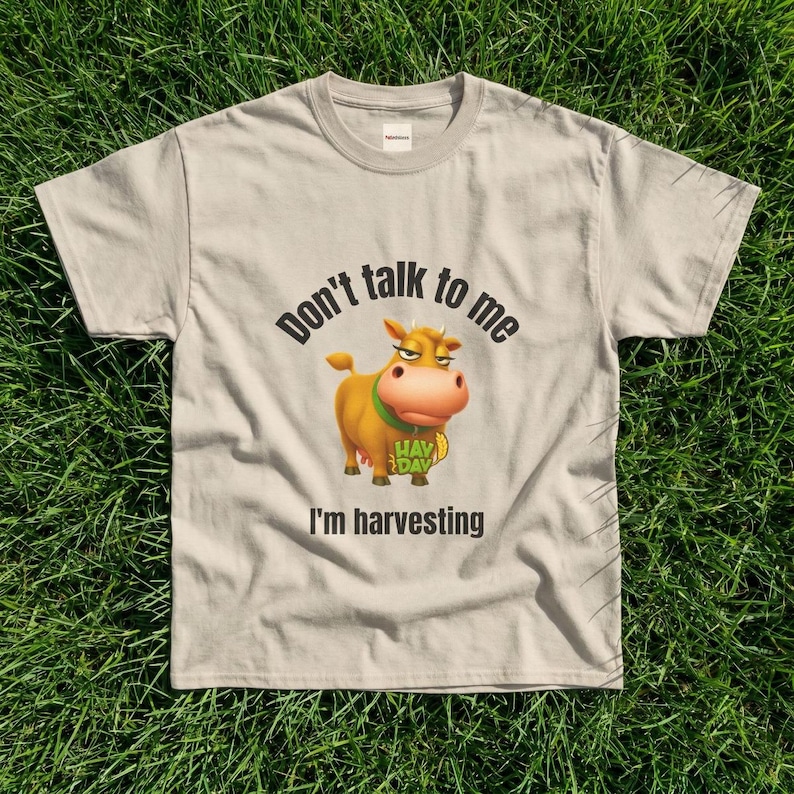 May include: A beige short-sleeved t-shirt featuring a graphic of a brown cow with a green collar. The text above the cow reads "Don't talk to me" and the text below reads "I'm harvesting". The cow also has the word "HAY DAY" printed on its chest.