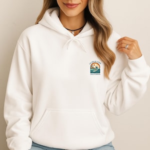 May include: White hooded sweatshirt with a front pocket and a small graphic on the upper left chest. The graphic features a surfer on a wave with the text "LA JOLLA COVE CALIFORNIA". The sweatshirt is a casual style.