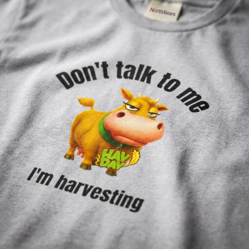 May include: A heather gray t-shirt featuring a cartoon cow with a green collar and the text "Don't talk to me I'm harvesting" printed in black. The graphic includes a yellow "HAY DAY" logo with a wheat stalk icon below the cow's chin.