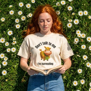 May include: A cream-colored short-sleeve t-shirt featuring a graphic of a brown cow holding a green bag with the text 'HAY DAY'. The shirt displays the black text 'Don't talk to me' above the cow and 'I'm harvesting' below the cow graphic.