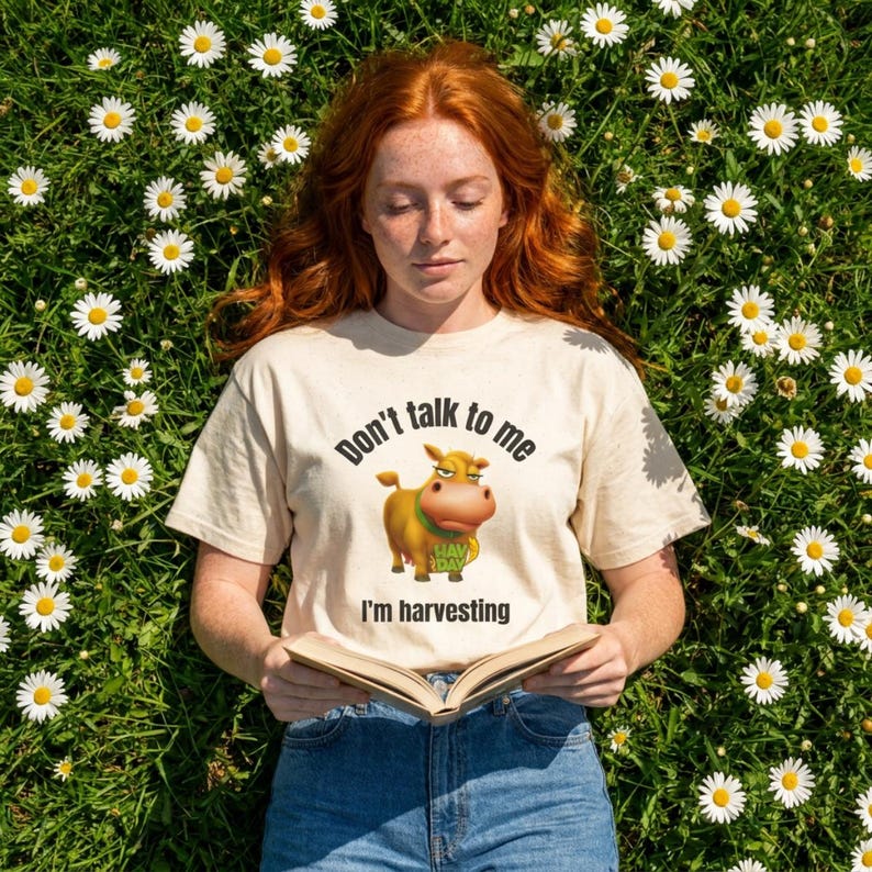 May include: A cream-colored short-sleeve t-shirt featuring a graphic of a brown cow holding a green bag with the text 'HAY DAY'. The shirt displays the black text 'Don't talk to me' above the cow and 'I'm harvesting' below the cow graphic.