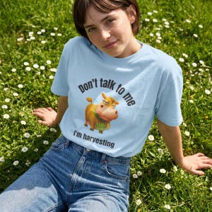 May include: A light blue t-shirt featuring a graphic of a cartoon cow with a green collar and the text "Don't talk to me I'm harvesting" printed in black. The cow is orange with a yellow snout and is holding a small bag labeled "HAY DAY".