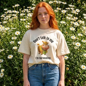 May include: A cream-colored short-sleeve t-shirt featuring a graphic of a brown cow with a green collar. The text above the cow reads Don't talk to me and the text below the cow reads I'm harvesting. The shirt is paired with blue denim jeans.