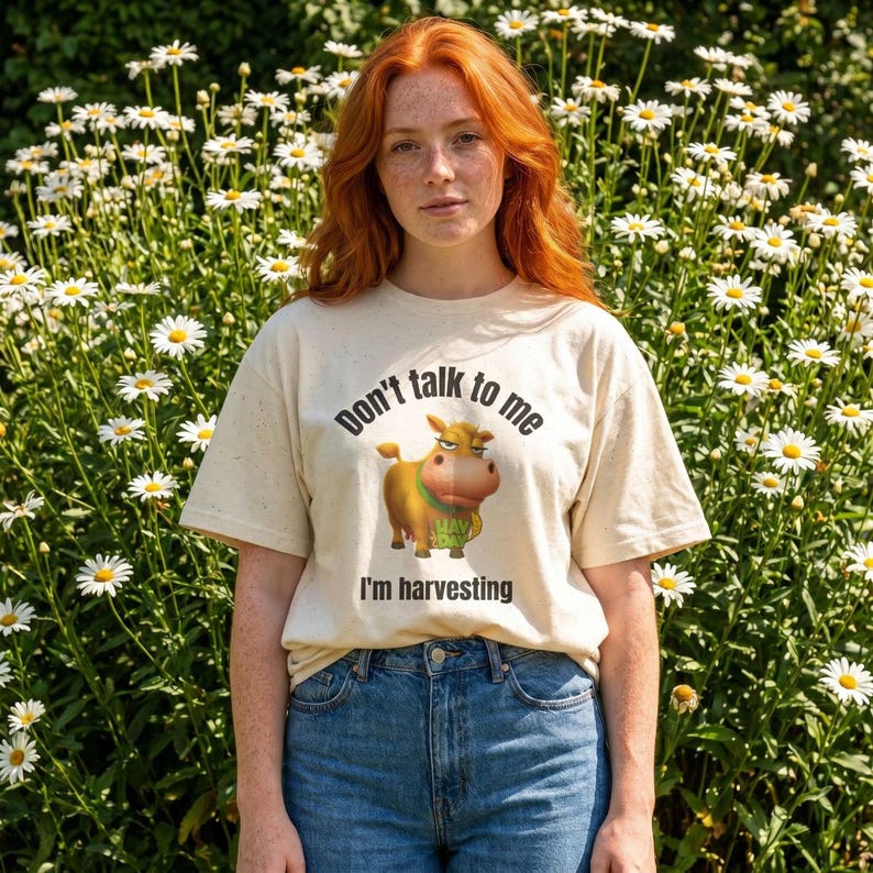 May include: A cream-colored short-sleeve t-shirt featuring a graphic of a brown cow with a green collar. The text above the cow reads Don't talk to me and the text below the cow reads I'm harvesting. The shirt is paired with blue denim jeans.