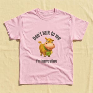May include: A light pink short-sleeved t-shirt featuring a cartoon cow graphic in the center. The cow wears a green collar and holds a wheat stalk. Above the cow is the text Don't talk to me and below is the text I'm harvesting.