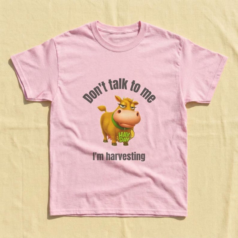 May include: A light pink short-sleeved t-shirt featuring a cartoon cow graphic in the center. The cow wears a green collar and holds a wheat stalk. Above the cow is the text Don't talk to me and below is the text I'm harvesting.