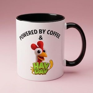 Puede incluir: Una taza de cerámica blanca con interior y asa negros. El frente presenta un gráfico de un pollo de dibujos animados sobre el texto verde "HAY DAY" y el texto negro "POWERED BY COFFEE &" arqueado sobre el pollo, todo sobre un fondo liso.