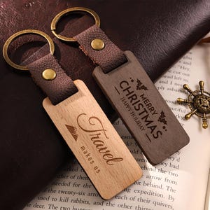 Puede incluir: Dos llaveros de madera con correas de cuero y anillos de latón. Un llavero es de color claro con "Travel makes us" grabado. El otro es marrón oscuro con "Merry Christmas Happy Holiday" grabado.