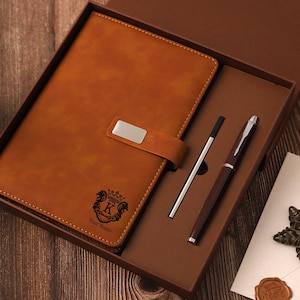 May include: A brown leather journal with a silver clasp and a matching pen and pencil set, presented in a gift box. The journal features a crest with the letter "K" and the words "Your Name". A star-shaped decoration and a wax seal are also included.